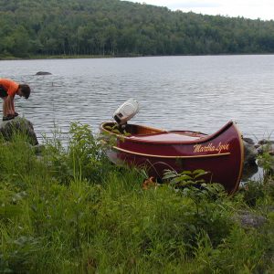 A boy ties his shoes next to a Northwoods-made Canoe on the bank of a river.